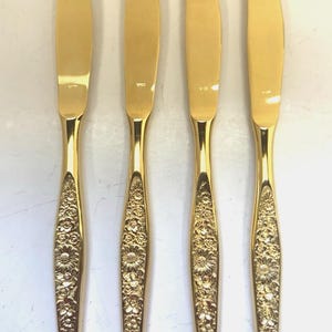May include: Four gold-colored butter knives with floral patterned handles. The blades are smooth and shiny, and the handles are intricately decorated with a repeating flower design. The knives are arranged vertically.