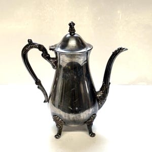 Puede incluir: Una tetera de plata con un asa y patas decorativas. La tetera tiene un acabado brillante y un diseño clásico.
