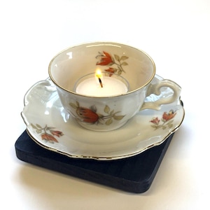 May include: A lit candle in a vintage teacup on a matching saucer. The white teacup and saucer have a gold trim and a floral design in orange and green. The teacup and saucer are on a black square base.