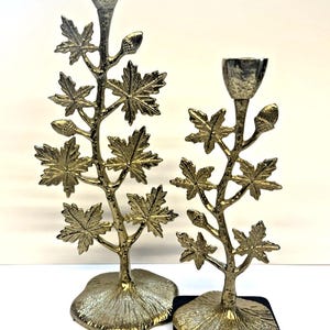 Puede incluir: Dos candelabros decorativos dorados con forma de ramas con hojas de arce. Uno se asienta sobre una base cuadrada negra, el otro sobre una base dorada con forma de hoja. Los candelabros tienen una copa plateada para sostener las velas.