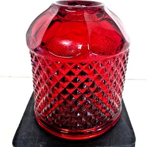 Puede incluir: Un tarro de vidrio rojo rubí con un patrón de diamantes, sobre una base cuadrada negra. El tarro tiene una parte superior redondeada y un cuerpo cilíndrico. El vidrio transparente permite el paso de la luz, resaltando el patrón.