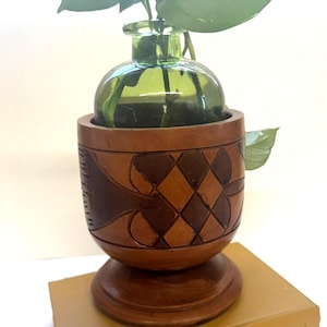 May include: A wooden planter with a carved design in shades of brown and a green glass bottle with a plant inside. The planter is sitting on a yellow book titled "The Green Thumb Garden Handbook".