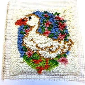 May include: A latch hook craft project featuring a white goose with an orange beak and black eye, surrounded by blue, green, and pink floral accents. The design is worked onto a square, off-white grid canvas with unfinished edges of mesh.