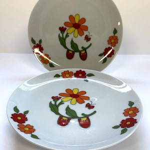 May include: Two white ceramic plates with a floral design. The plates feature a central image of a flower with orange, yellow, and green details, and red shoes. Additional flowers in red and orange are placed around the edges.