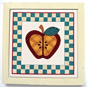 Op de afbeelding: Vierkante decoratieve tegel met een crèmekleurige lijst. De tegel heeft een gestileerd appelontwerp met een geel en rood ruitpatroon, een bruine steel en een turquoise blad met zwarte stippen. De rand heeft een turquoise en crème dambordpatroon.