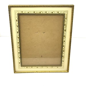 May include: A rectangular picture frame with a gold-colored metal frame and a cream-colored border. The border features a repeating pattern of green and black leaf-like shapes. The frame has a cardboard backing and a glass front.