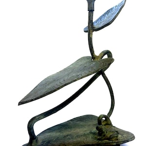 May include: A bronze-effect candle holder with a botanical design. It features a central stem with a candle cup, supported by stylized leaves. The base is a dark, rectangular shape. The design evokes nature, with leaf-like forms.
