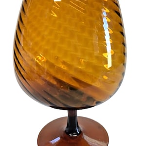 Może przedstawiać: Bursztynowy, ręcznie dmuchany kieliszek do brandy. Czarka ma teksturowany, wirujący wzór, a nóżka i podstawa są ciemnobrązowe. Szkło jest przezroczyste. Kieliszek przeznaczony jest do serwowania brandy.