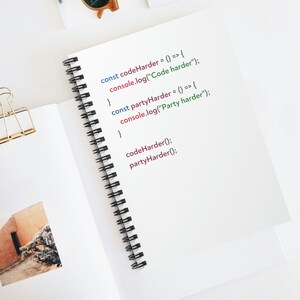 May include: A spiral bound notebook with handwritten code on the pages. The code includes the text "const codeHarder = () => { console.log("Code harder"); }" and "const partyHarder = () => { console.log("Party harder"); }" and the lines "codeHarder();" and "partyHarder();".