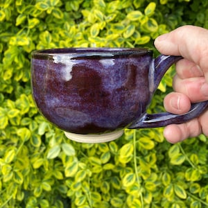 Puede incluir: Una taza de cerámica hecha a mano con un esmalte morado oscuro. La taza tiene una forma redondeada, un asa robusta y una base clara. El fondo es un follaje verde y amarillo vibrante.