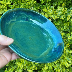 Op de afbeelding: Een ronde keramische kom met een glanzend glazuur in gemengde tinten van diep turkoois, smaragdgroen en vleugjes donkerblauw. Het oppervlak heeft een gladde, reflecterende afwerking met subtiele kleurvariaties in de kom zelf.