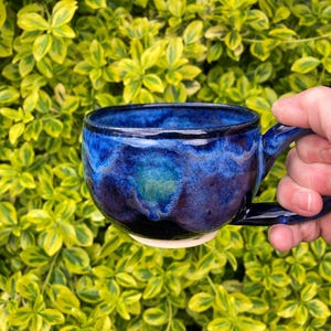 Puede incluir: Una taza de cerámica hecha a mano con un esmalte azul oscuro, con tonos índigo y verde azulado. La taza tiene una forma redondeada con un asa y una base blanca. El fondo es una vibrante planta de hojas verdes y amarillas.