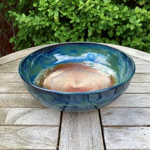 May include: A round ceramic bowl featuring a glossy glaze with a deep blue and green exterior. The interior of the bowl transitions from a dark blue rim to a warm, earthy copper-brown centre with a smooth, reflective surface finish.