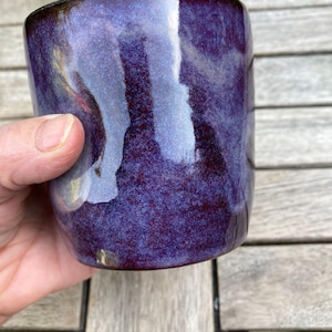 May include: A cylindrical ceramic mug featuring a glossy, mottled glaze in deep shades of purple, violet, and dark blue. The surface has a smooth, reflective finish with subtle variations in colour intensity and a small light-coloured speck near the base.