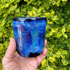 May include: A ceramic tumbler featuring a glossy glaze with deep blue, cobalt, and black tones. The surface displays a smooth, mottled texture with blended colour transitions that flow from the rim down to the rounded base of the vessel.
