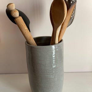 Może przedstawiać: Szary ceramiczny pojemnik na przybory kuchenne wypełniony drewnianymi łyżkami i czarną szpatułką. Łyżki mają różne kolory, od jasnobrązowego do ciemnobrązowego. Pojemnik ma gładkie, błyszczące wykończenie i cylindryczny kształt. Przybory są ustawione pionowo.