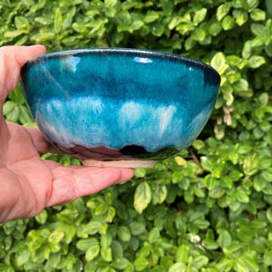 May include: A ceramic bowl featuring a glossy glaze that transitions from a deep, dark teal at the rim to a lighter, vibrant turquoise in the centre, ending in a dark brown base. The bowl has a smooth, rounded shape with a visible unglazed clay foot.