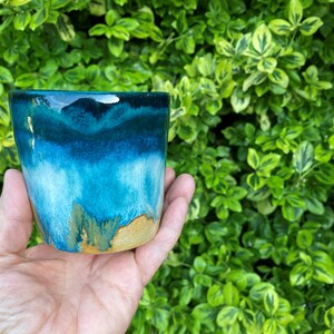 May include: A ceramic cup featuring a reactive glaze with a deep navy blue rim that transitions into a vibrant turquoise and light blue body, ending in a raw, unglazed tan clay base with visible drips and organic colour blending throughout.