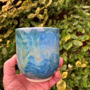 Op de afbeelding: Een handgemaakte keramische beker met een blauw en turquoise gemarmerde glazuur. De beker heeft een afgeronde vorm en een lichtbeige basis. De achtergrond toont groen en geel blad.