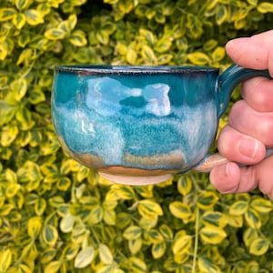 Op de afbeelding: Een handgemaakte keramische mok met een turquoise glazuur. De mok heeft een afgeronde vorm, een oor en een bruine basis. De turquoise kleur vloeit over in een lichter blauw, waardoor een gradiënteffect ontstaat. De mok wordt vastgehouden tegen een achtergrond van groen en geel blad.