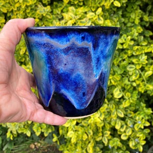 May include: A handcrafted ceramic cup with a glossy, deep blue glaze. The cup's interior and exterior have a marbled effect with shades of blue, purple, and black. The cup is held in a hand against a backdrop of green and yellow foliage.