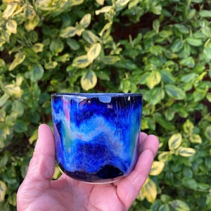Può includere: Una piccola tazza in ceramica con una smaltatura lucida caratterizzata da motivi vorticosi di blu reale profondo, turchese vibrante e tocchi di nero. Il recipiente cilindrico presenta un bordo liscio e arrotondato e una base leggermente affusolata.