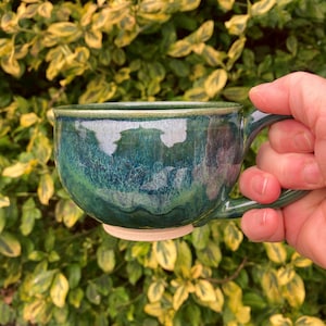 May include: A handmade ceramic mug with a deep teal glaze. The mug has a rounded body, a sturdy handle, and a light beige base. The glaze has a glossy finish with variations in colour, creating a unique, artistic look. The mug is held in a hand.