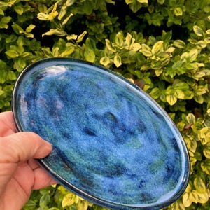 Könnte beinhalten: Ovale Keramik-Servierplatte mit glänzender, marmorierter blau-grüner Glasur. Die Platte wird vor einem Hintergrund aus grünem und gelbem Laub gehalten. Der Rand der Platte ist dunkler blau.