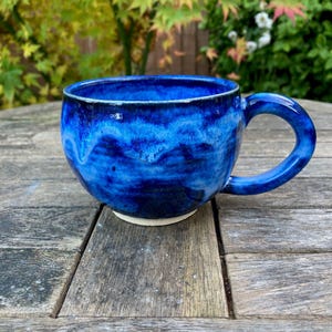 Könnte beinhalten: Handgefertigte Keramik-Tasse mit tiefblauer, glänzender Glasur. Die Tasse hat eine runde Form, einen stabilen Henkel und einen weißen Boden. Die blaue Glasur hat einen Marmoreffekt, der ein einzigartiges Muster erzeugt. Ideal für Kaffee oder Tee.