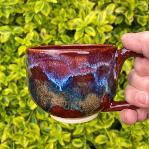 Può includere: Una tazza in ceramica fatta a mano con una glassa lucida. La tazza presenta una miscela di tonalità rosso scuro, blu e marrone, creando un effetto marmorizzato unico. Ha una forma arrotondata, un manico robusto e una base bianca. Lo sfondo è verde vibrante.