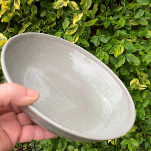 May include: A pale grey, oval ceramic bowl with a textured surface and a pinkish rim. The bowl is set against a backdrop of green and yellow foliage. The image suggests a handmade or artisan product.
