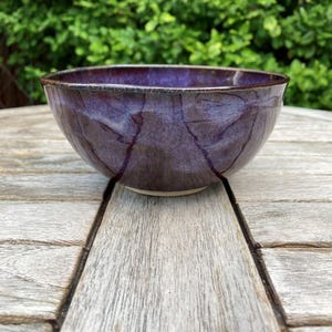 May include: A round ceramic bowl featuring a glossy, deep purple glaze with subtle, darker veining patterns throughout the surface. The bowl has a smooth, curved silhouette and a dark, defined rim, showcasing a handcrafted finish with a light-coloured base.