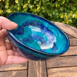 May include: A handcrafted ceramic bowl with a striking, swirling design in shades of turquoise, teal, and deep blue. The bowl has a smooth, glossy surface and an oval form. The bowl is held in a hand, highlighting its colours and design.