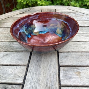 May include: A round ceramic bowl with a glossy finish, featuring a deep reddish-brown base colour blended with vibrant streaks of blue and gold glaze. The interior of the bowl displays a smooth, reflective surface with swirling colour patterns.