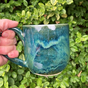 May include: A ceramic mug featuring a glossy, multi-tonal glaze in shades of deep forest green, teal, and bright emerald. The surface displays a fluid, blended colour pattern with a smooth, reflective finish and a curved handle on the side.