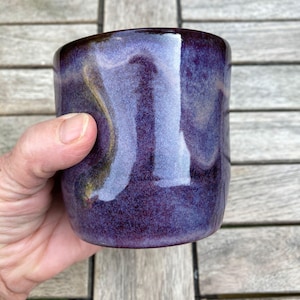 Puede incluir: Una taza de cerámica hecha a mano con un esmalte morado oscuro. La taza tiene una forma redondeada y un borde marrón oscuro. El esmalte tiene un acabado brillante, que refleja la luz y crea una textura visual única. La taza se sostiene en una mano.