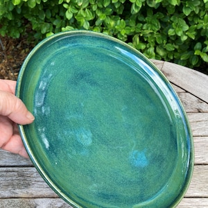 Puede incluir: Un plato ovalado de cerámica con un esmalte brillante y matizado en tonos verde bosque profundo y azul cerceta. La superficie presenta sutiles variaciones de color y un acabado liso y reflectante con un borde ligeramente elevado.