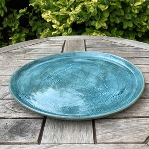 Puede incluir: Un plato redondo de cerámica turquesa con una superficie texturizada. El plato se exhibe sobre una mesa de madera, con follaje verde en el fondo. El plato tiene aproximadamente 30 cm de diámetro.