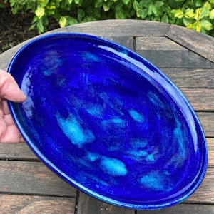 May include: An oval ceramic platter in a striking, deep blue glaze with lighter blue marbling. The dish is presented on a wooden table. The platter's dimensions are suitable for serving food, adding a touch of elegance to any table setting.