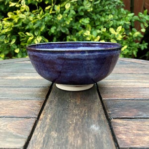 Può includere: Una ciotola in ceramica fatta a mano con una profonda glassa indaco. La ciotola ha una forma arrotondata con un bordo leggermente svasato e una base di colore chiaro. La ciotola è su una superficie di legno, con fogliame verde sullo sfondo. Ideale per servire zuppe, insalate o snack.