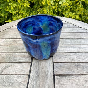 Peut inclure: Un bol en céramique fait à la main avec un glaçage bleu profond, présentant des stries turquoise et noires. Le bol a une forme organique unique et repose sur une table en bois. L'arrière-plan est une vue floue du feuillage vert.