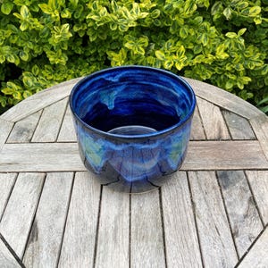 May include: A round ceramic planter featuring a deep blue glaze with swirling lighter blue and subtle green accents. The vessel has a smooth, glossy finish and a wide, open top, showcasing a rich, multi-tonal colour gradient across its exterior surface.