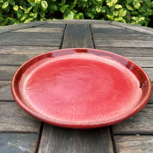 May include: A circular, red ceramic plate with a glossy finish. The plate has a slightly raised edge and a smooth surface. The plate is on a wooden table.