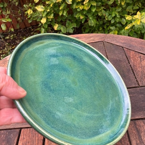 May include: A handmade, oval ceramic plate with a glossy, mottled green glaze. The plate has a slightly raised rim and a smooth surface. The plate is held by a hand, and the background is a wooden table and foliage.