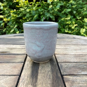 Puede incluir: Un vaso de cerámica hecho a mano con un esmalte moteado azul-gris apagado. La taza cilíndrica tiene una base redondeada y un borde liso. La taza está sobre una mesa de madera, con follaje verde en el fondo. La taza mide aproximadamente 10 cm de alto.
