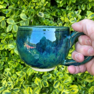 Könnte beinhalten: Ein Keramikbecher mit glänzender Oberfläche in einer Mischung aus tiefblauer und leuchtend grüner Glasur. Der Becher hat einen abgerundeten Körper, einen stabilen Henkel und einen glatten, unglasierten hellen Boden. Die Oberfläche reflektiert Licht.