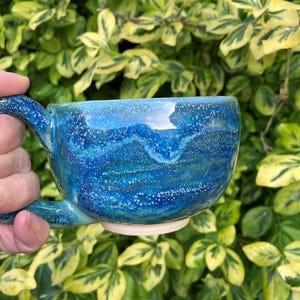 May include: A handcrafted ceramic mug with a striking blue glaze, speckled with white. The mug has an unusual, organic form and a robust handle. The background shows green and yellow foliage.