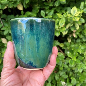 May include: A ceramic cup featuring a glossy, reactive glaze with blended shades of deep forest green, teal, and navy blue. The vessel has a smooth, cylindrical shape with a slightly tapered base and a visible light-coloured clay rim at the bottom.