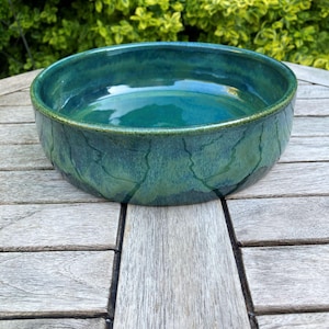 May include: A teal-green glazed ceramic bowl. The bowl has a wide, shallow shape and a textured surface. The rim is a darker shade of green. The bowl is sitting on a wooden table. The bowl's diameter is approximately 25 cm.