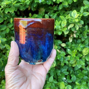 May include: A cylindrical ceramic cup featuring a glossy glaze with a deep brown rim that transitions into a vibrant, mottled blue and teal body. The surface has a smooth, reflective finish with subtle color variations throughout the vessel.